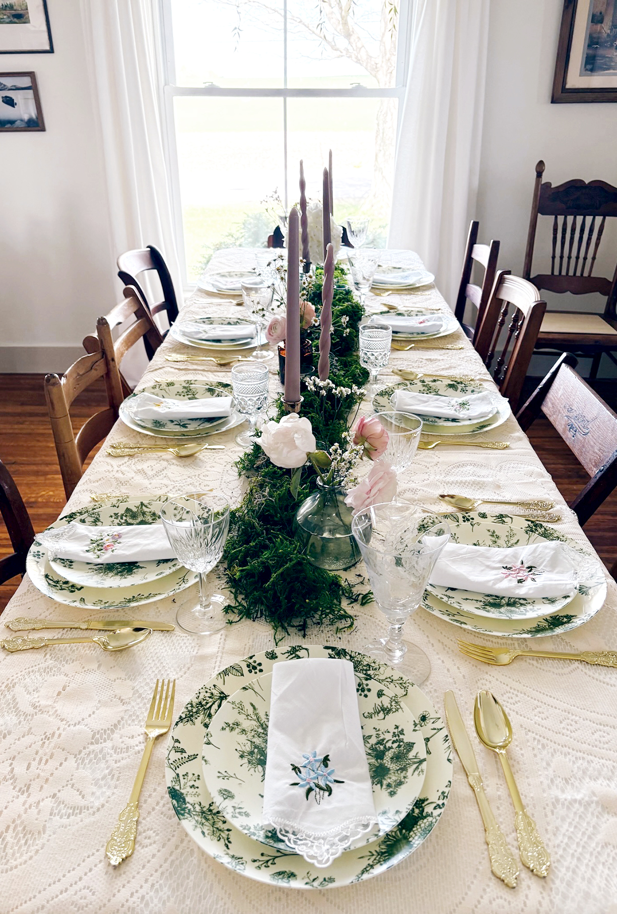table with moss centerpiece and taupe candles and fresh flowers with place settings around it with green floral plates and embroidered napkins on plates